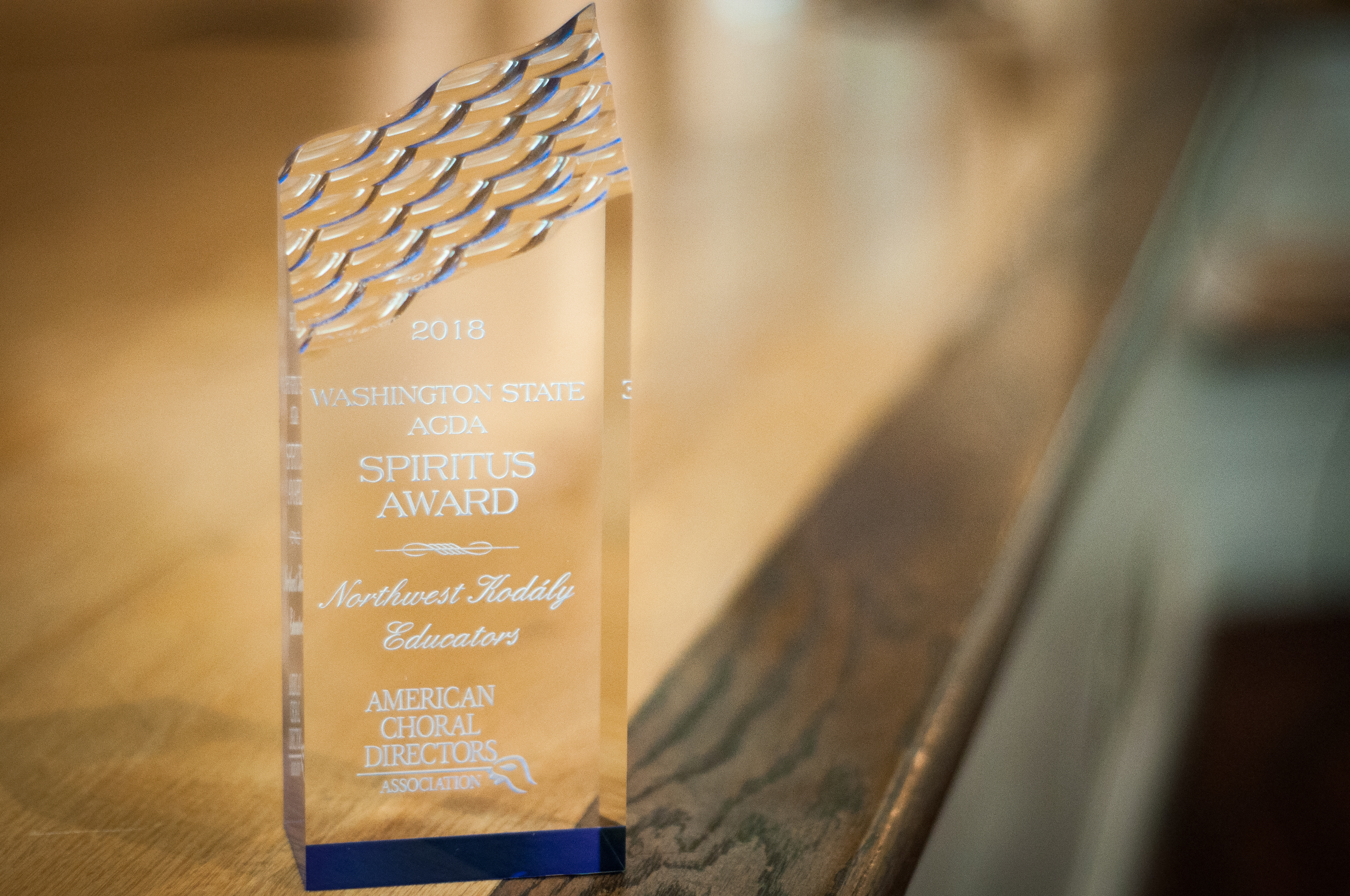This image is a picture of the Spiritus Award given to NWKE by Washington State ACDA for our significant contribution to choral music in Washington State. The award is a glass prism with an angled top that has a wavy texture. The text on the award reads "2018 Washington State ACDA Spiritus Award - Northwest Kodály Educators - American Choral Directors Association."